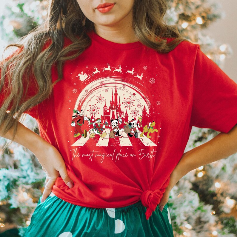 Puede incluir: Camiseta roja con un gr&aacute;fico blanco de Mickey Mouse y sus amigos caminando por un paso de cebra frente al Castillo de Cenicienta. El gr&aacute;fico tambi&eacute;n incluye copos de nieve, fuegos artificiales y el texto "The most magical place on Earth".
