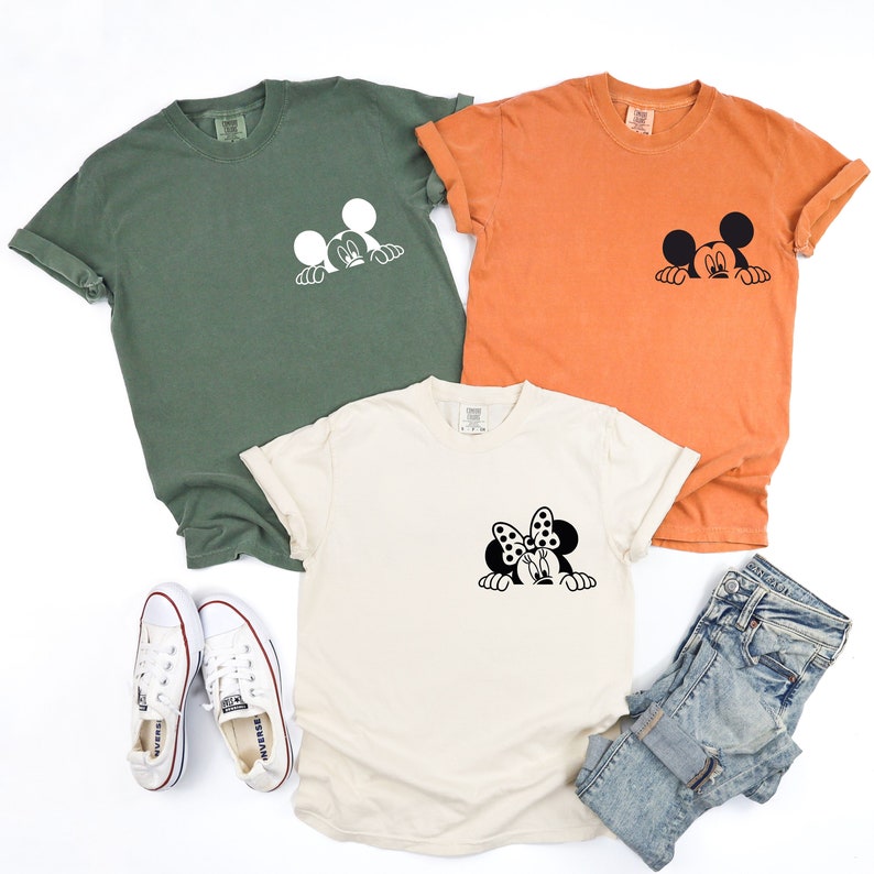 May include: Three t-shirts with a Mickey Mouse and Minnie Mouse peeking design. The shirts are in olive green, orange, and cream colors.