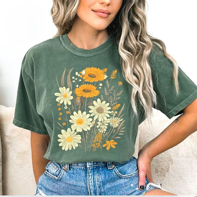 May include: Green t-shirt featuring a floral design with yellow and white daisies and sunflowers. The graphic includes various wildflowers and foliage in shades of yellow and brown. The shirt is made of a soft material.