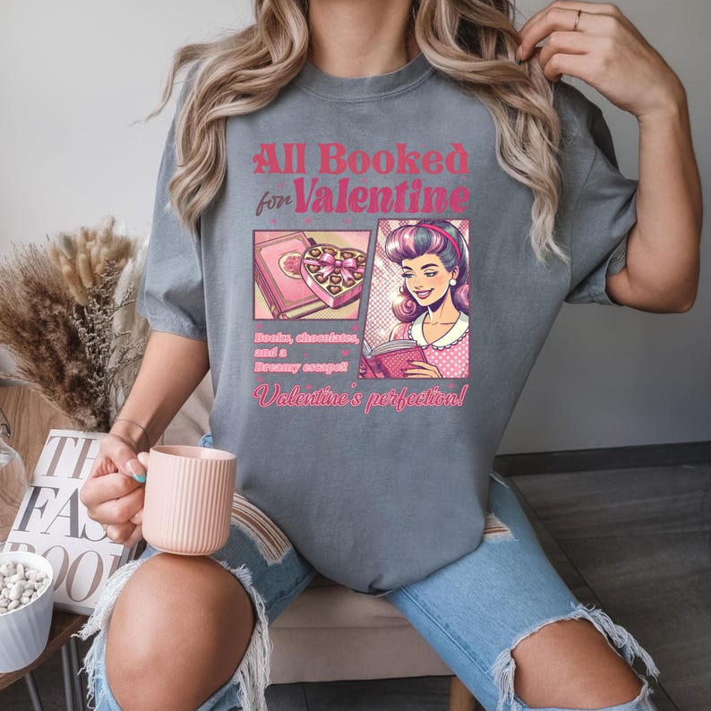 Puede incluir: Camiseta gris con un dise&ntilde;o gr&aacute;fico que presenta a una mujer leyendo un libro y una caja de chocolates. El texto en la camiseta dice "All Booked for Valentine" y "Books, chocolate, and a dreamy escape! Valentine's perfection!"