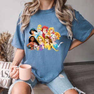 Puede incluir: Una camiseta azul con un estampado gr&aacute;fico de princesas Disney, incluyendo Cenicienta, Ariel, Bella y Moana.
