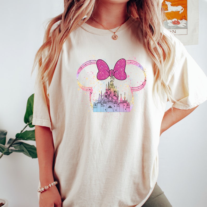 Puede incluir: Una camiseta blanca con un lazo rosa y una ilustraci&oacute;n colorida de un castillo. El castillo est&aacute; rodeado de un efecto acuarela azul claro, rosa y amarillo.