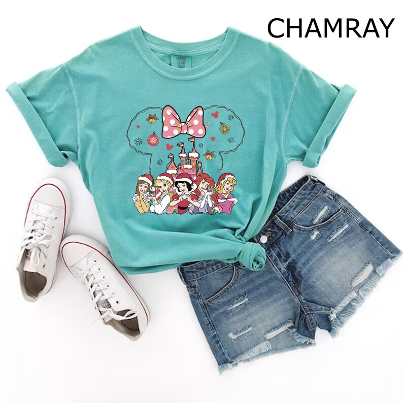 Puede incluir: Una camiseta azul turquesa con un gr&aacute;fico navide&ntilde;o de princesas Disney frente a un castillo. El gr&aacute;fico incluye un lazo rojo y adornos. La camiseta se combina con pantalones cortos de mezclilla y zapatillas blancas.