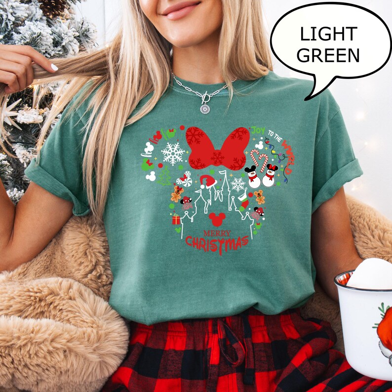 K&ouml;nnte beinhalten: Hellgr&uuml;nes T-Shirt mit einem weihnachtlichen Minnie-Mouse-Design. Das Design umfasst eine rote Schleife, Schneeflocken und die Worte "Merry Christmas". Das Shirt ist ein l&auml;ssiger, bequemer Stil.