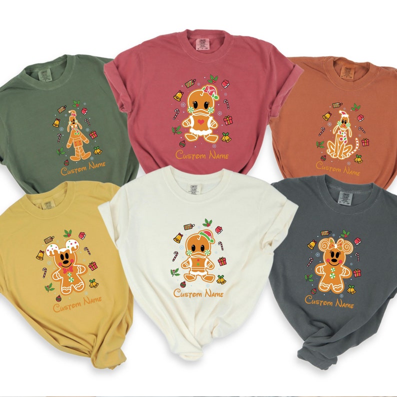 May include: Six different colored t-shirts with a gingerbread character design. Each shirt has a different character, including Mickey Mouse, Donald Duck, and Pluto. The shirts are all in festive holiday colors and have the text "Custom Name" printed on the bottom.