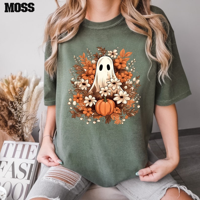 Op de afbeelding: Een groen T-shirt met een witte geest omringd door oranje en witte bloemen en een kleine oranje pompoen. De geest heeft twee zwarte ogen.