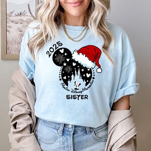 Peut inclure: T-shirt bleu clair avec une silhouette noire et blanche de Mickey Mouse portant un bonnet de P&egrave;re No&euml;l. La silhouette comprend le texte "2025", "Making Disney Memories", et "Sister".