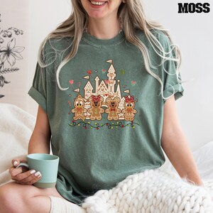 May include: A moss green t-shirt featuring a festive graphic. The graphic includes a castle, gingerbread characters, and holiday lights. The word "MOSS" is printed in black at the top right corner of the shirt.