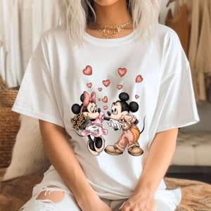 May include: White t-shirt with a graphic of Mickey Mouse and Minnie Mouse holding hands with hearts floating around them.