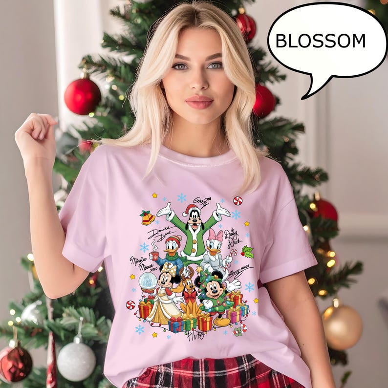 Puede incluir: Camiseta rosa claro con un dise&ntilde;o navide&ntilde;o de Disney. El gr&aacute;fico incluye a Mickey Mouse, Minnie Mouse, Donald Duck, Daisy Duck y Goofy, rodeados de regalos, copos de nieve y bastones de caramelo. La palabra "Blossom" est&aacute; en una burbuja.