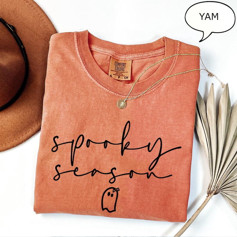 May include: A coral-coloured Comfort Colors t-shirt with the words "spooky season" in black script and a ghost graphic. A gold necklace and a brown hat are also visible. The word "YAM" is in a speech bubble.
