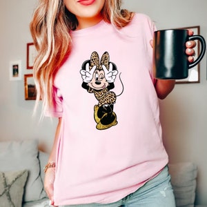 K&ouml;nnte beinhalten: Hellrosa T-Shirt mit einer Cartoon-Figur von Minnie Mouse in einem Leopardenmuster-Outfit. Minnie macht mit ihren H&auml;nden ein Friedenszeichen. Die Schuhe der Figur sind goldfarben, und ihre Schleife und ihr Outfit haben ein Leopardenmuster.