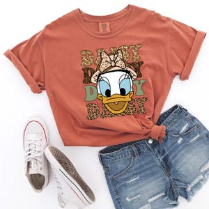 May include: A rust-coloured t-shirt featuring a cartoon image of Daisy Duck wearing a bow and a hat. The text "Daisy Daisy Daisy" is printed on the shirt in a retro font with animal print accents.