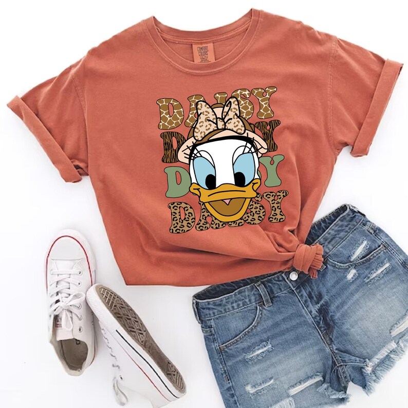 May include: A rust-coloured t-shirt featuring a cartoon image of Daisy Duck wearing a bow and a hat. The text "Daisy Daisy Daisy" is printed on the shirt in a retro font with animal print accents.