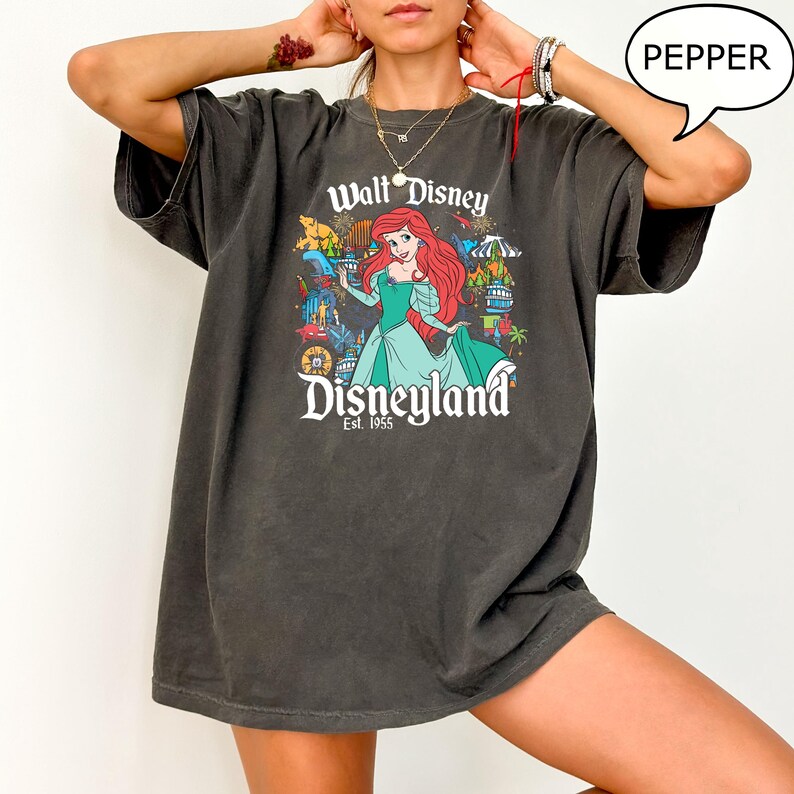 May include: Dark grey t-shirt with a graphic of Ariel from Disney's The Little Mermaid, and the text "Walt Disney Disneyland Est. 1955". The design includes various park attractions and characters. The shirt has a relaxed fit.