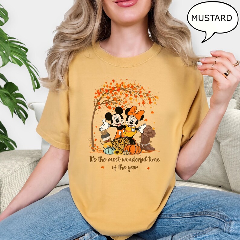 May include: Mustard-colored t-shirt featuring Mickey and Minnie Mouse under a tree with autumn leaves. The graphic includes pumpkins, a boot, and the text "It's the most wonderful time of the year."