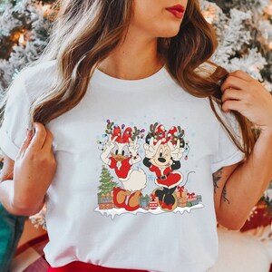 Puede incluir: Camiseta blanca con un gr&aacute;fico navide&ntilde;o con Minnie Mouse y Daisy Duck con gorros de Pap&aacute; Noel y cuernos de reno. El gr&aacute;fico incluye un &aacute;rbol de Navidad, regalos y copos de nieve.