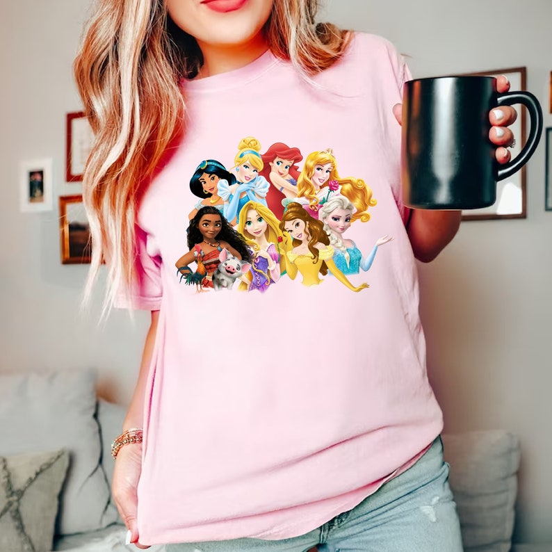 Puede incluir: Camiseta rosa con un gr&aacute;fico de princesas Disney, incluyendo Cenicienta, Ariel, Bella y Moana.