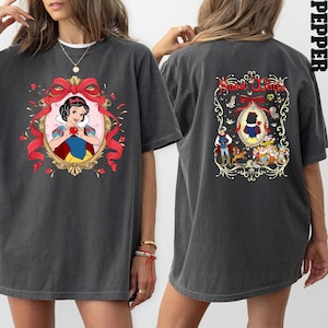 May include: Dark gray t-shirt featuring a Snow White graphic on the front and back. The front design includes a portrait of Snow White holding an apple within a decorative frame. The back design features the text "Snow White" and additional characters.