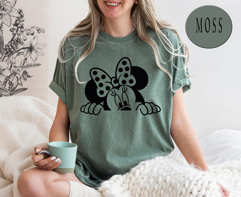 K&ouml;nnte beinhalten: Ein gr&uuml;nes T-Shirt mit einer schwarzen und wei&szlig;en Grafik von Minnie Mouse, die &uuml;ber den Rand schaut. Der Text "MOSS" befindet sich in einem gr&uuml;nen Kreis in der oberen rechten Ecke des Bildes.