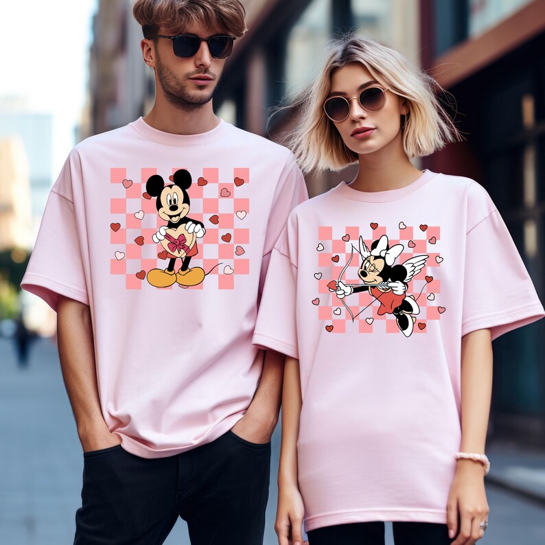 Puede incluir: Camisetas rosas con dise&ntilde;os de Mickey y Minnie Mouse. Una camiseta tiene a Mickey sosteniendo un regalo en forma de coraz&oacute;n, la otra tiene a Minnie como Cupido con arco y flecha. Ambas camisetas tienen un fondo a cuadros rosa y blanco con corazones.
