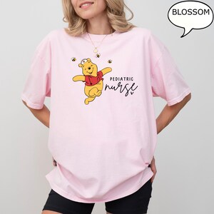 May include: A light pink t-shirt featuring a cartoon bear in a red shirt and nurse hat, with the words "Pediatric Nurse" in black script. Small bee graphics are scattered around the bear. The word "Blossom" is in a speech bubble.