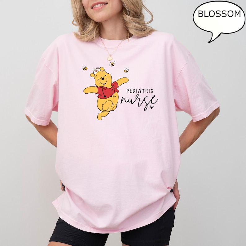 May include: A light pink t-shirt featuring a cartoon bear in a red shirt and nurse hat, with the words "Pediatric Nurse" in black script. Small bee graphics are scattered around the bear. The word "Blossom" is in a speech bubble.