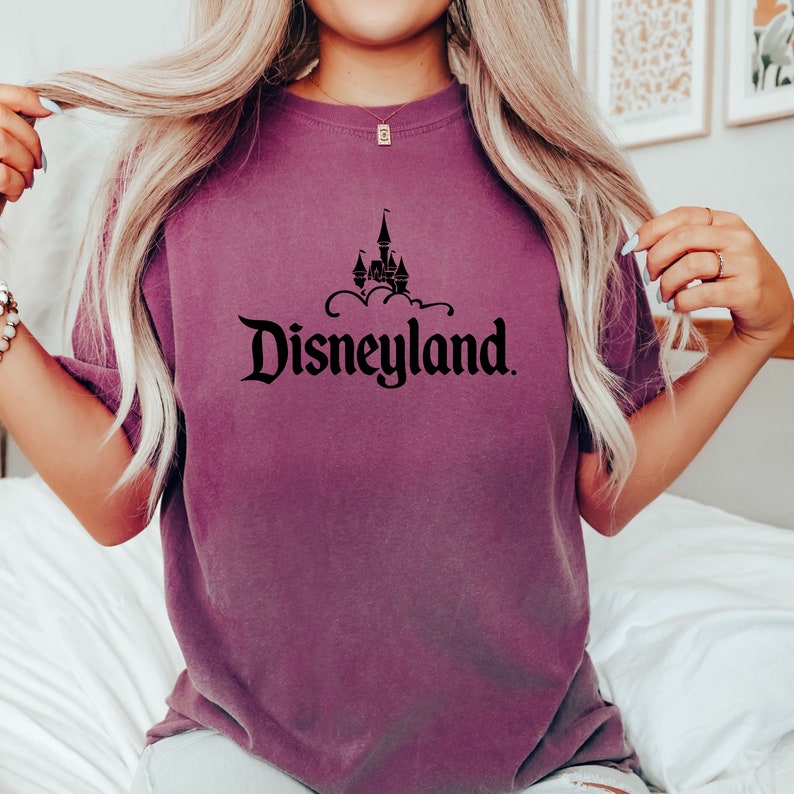 Puede incluir: Camiseta malva con la palabra "Disneyland" en fuente negra y un castillo como gr&aacute;fico. La camiseta es de color liso y tiene un corte holgado. El dise&ntilde;o est&aacute; centrado.