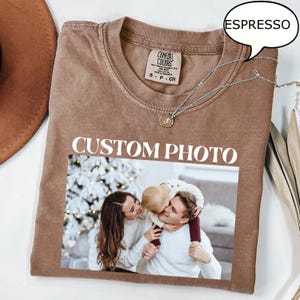 May include: Espresso-colored t-shirt with a photo print. The shirt features the text "CUSTOM PHOTO" in white block letters. The photo shows a family of three. A silver necklace with a pendant is draped on the shirt.