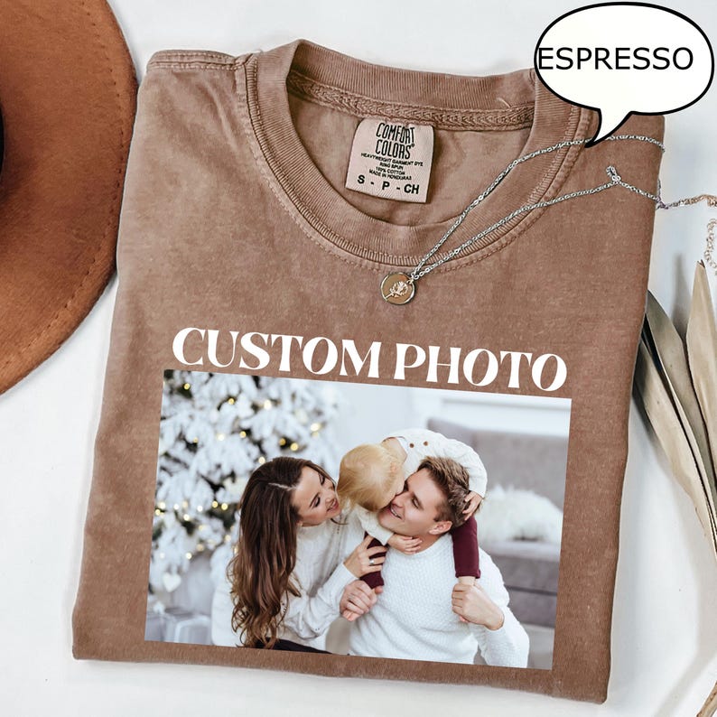 May include: Espresso-colored t-shirt with a photo print. The shirt features the text "CUSTOM PHOTO" in white block letters. The photo shows a family of three. A silver necklace with a pendant is draped on the shirt.