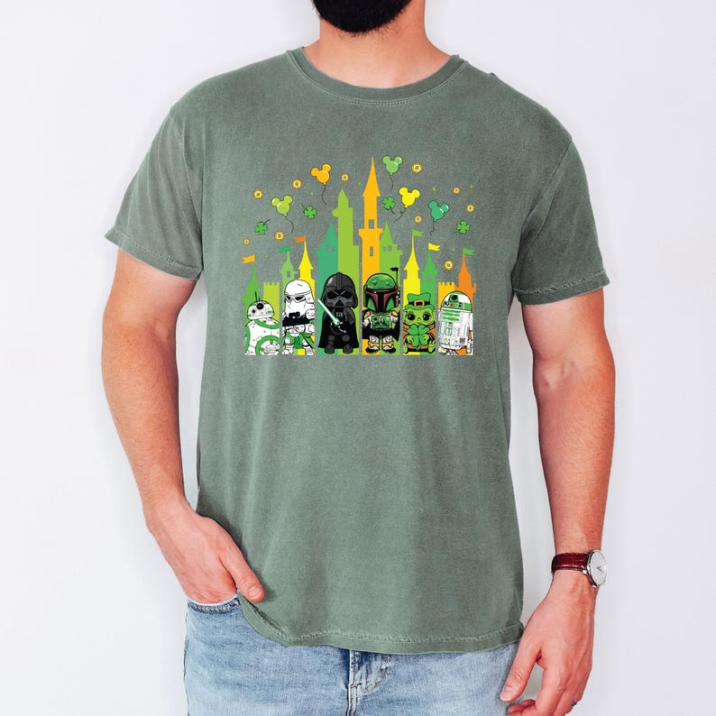 Puede incluir: Camiseta verde con un dise&ntilde;o colorido de personajes de Star Wars frente a un castillo. El dise&ntilde;o incluye a R2-D2, Darth Vader y otros personajes, con tr&eacute;boles y orejas de Mickey Mouse. La camiseta es de material suave.