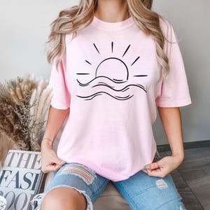 May include: A pink t-shirt with a black graphic of a sun setting over waves. The graphic is simple and minimalist.