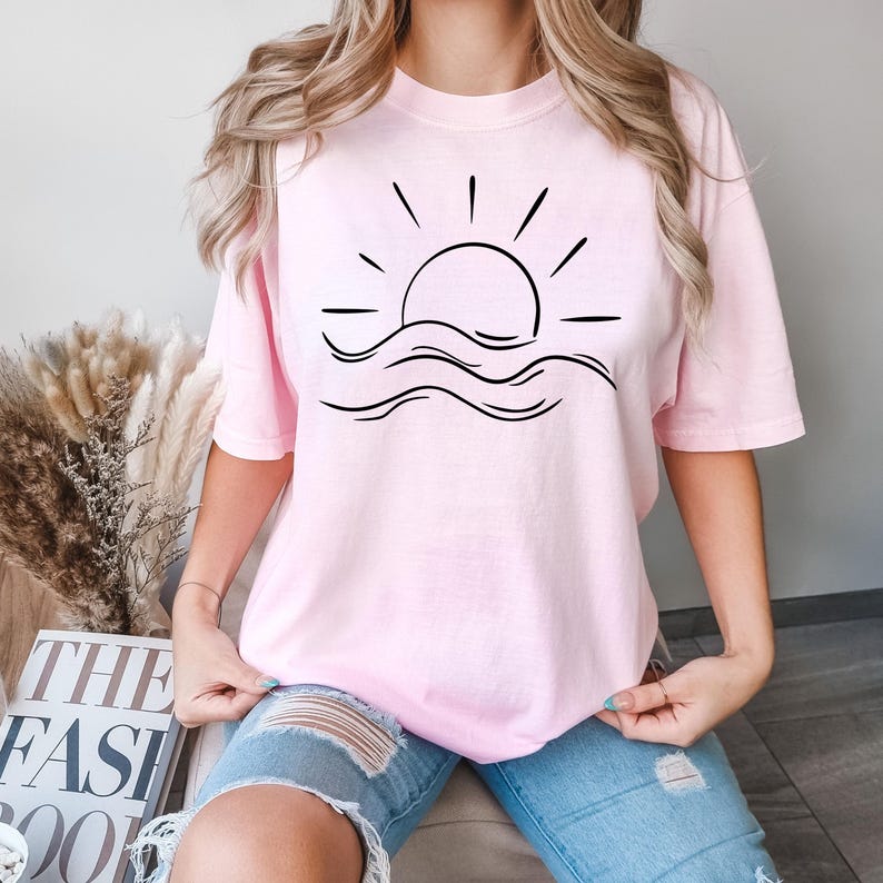 May include: A pink t-shirt with a black graphic of a sun setting over waves. The graphic is simple and minimalist.