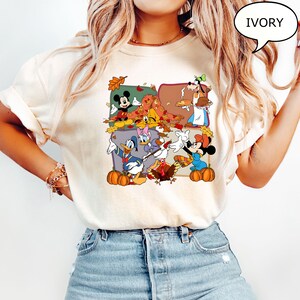 May include: Ivory-colored t-shirt featuring a colorful graphic of Disney characters celebrating autumn. Mickey Mouse, Minnie Mouse, Donald Duck, and Goofy are surrounded by pumpkins and fall leaves. The word "IVORY" is in a speech bubble.