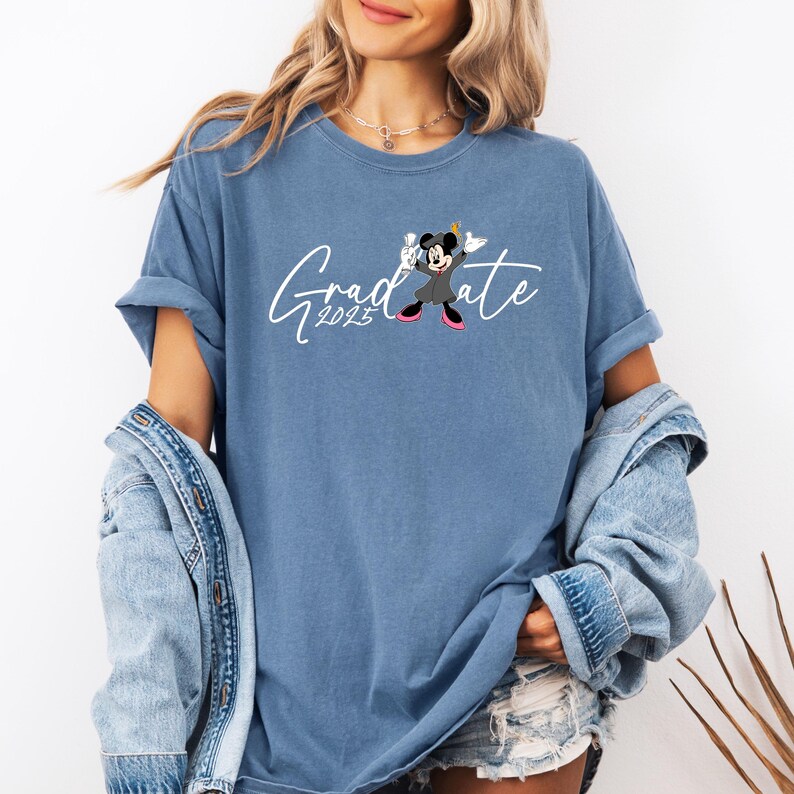 Puede incluir: Una camiseta azul con un gr&aacute;fico blanco de Mickey Mouse vestido con una gorra y una bata de graduaci&oacute;n. El texto "Graduate 2023" est&aacute; escrito en letras blancas en cursiva.