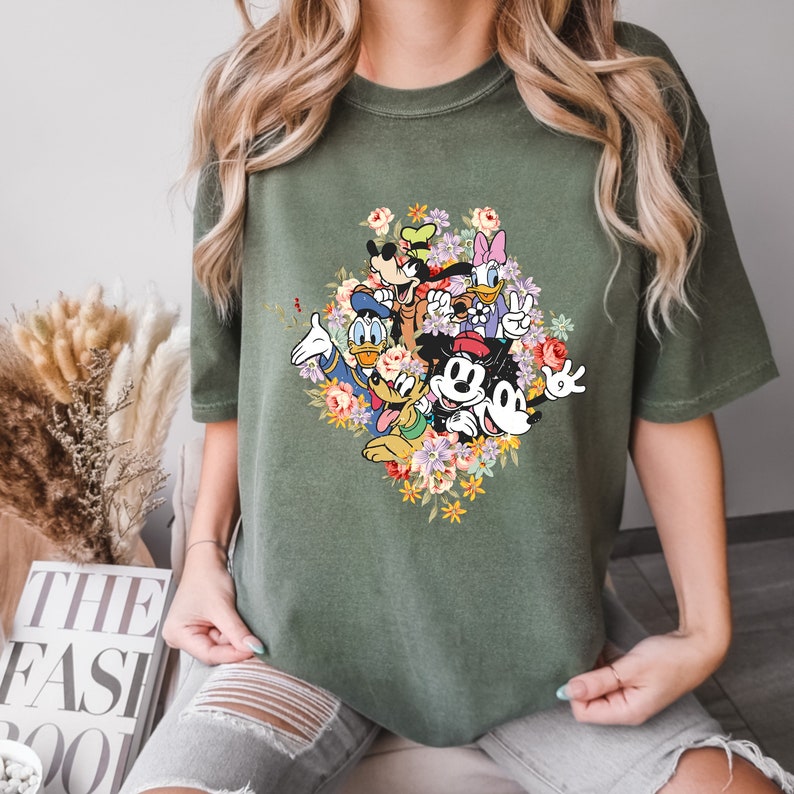 May include: A green t-shirt featuring a colourful graphic print of Mickey Mouse and his friends surrounded by flowers.