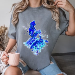 Puede incluir: Una camiseta gris con una ilustraci&oacute;n de acuarela azul de una sirena sentada en una roca. La sirena est&aacute; silueteada sobre un fondo azul y blanco.
