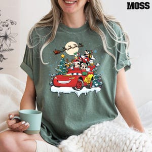 May include: A green t-shirt with a Christmas-themed design featuring Mickey Mouse, Donald Duck, Goofy, and Pluto riding in a red Lightning McQueen car. The characters are wearing Santa hats and there are Christmas trees in the background.