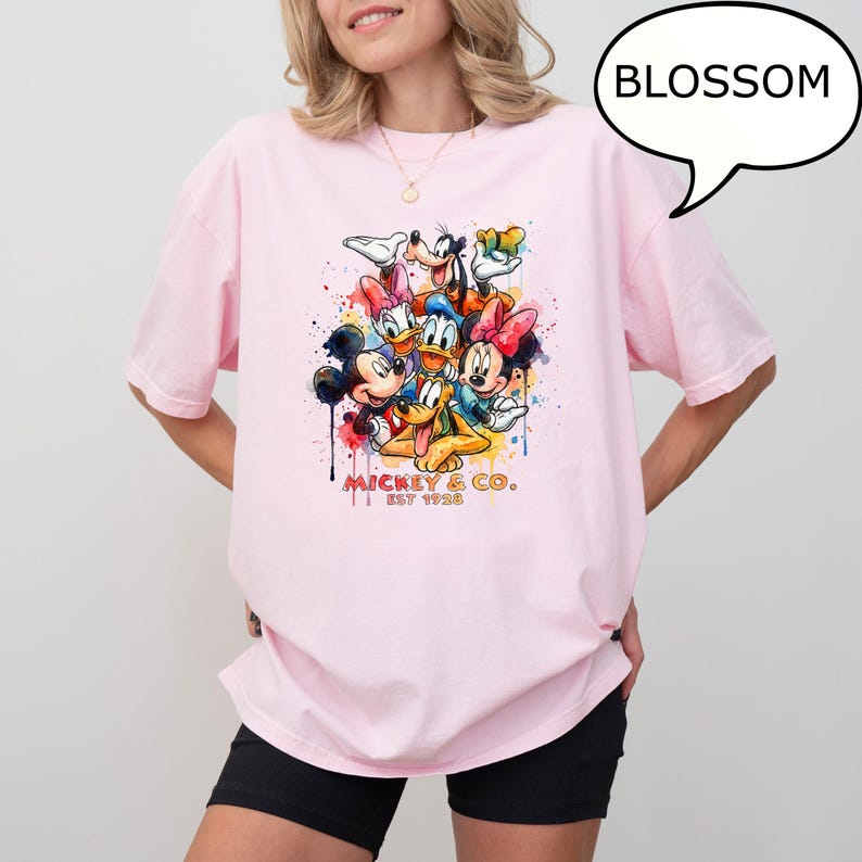 May include: A light pink t-shirt featuring a colorful watercolor graphic of Mickey Mouse, Minnie Mouse, Donald Duck, Daisy Duck, Goofy, and Pluto. The text "Mickey & Co. Est 1928" is printed below the characters. A speech bubble with the word "BLOSSOM" is in the upper right.