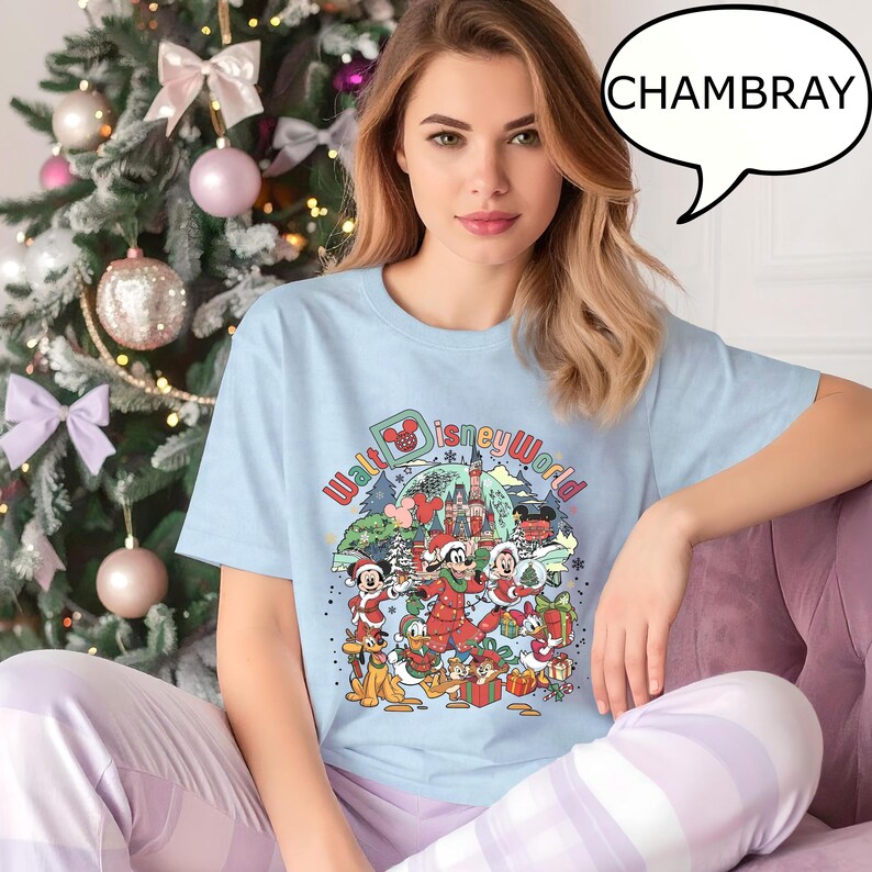 May include: Light blue t-shirt with a colorful graphic of Disney characters in a winter scene, including Mickey Mouse, Minnie Mouse, and Donald Duck. The text "Walt Disney World" is also visible. The word "CHAMBRAY" is in a speech bubble.