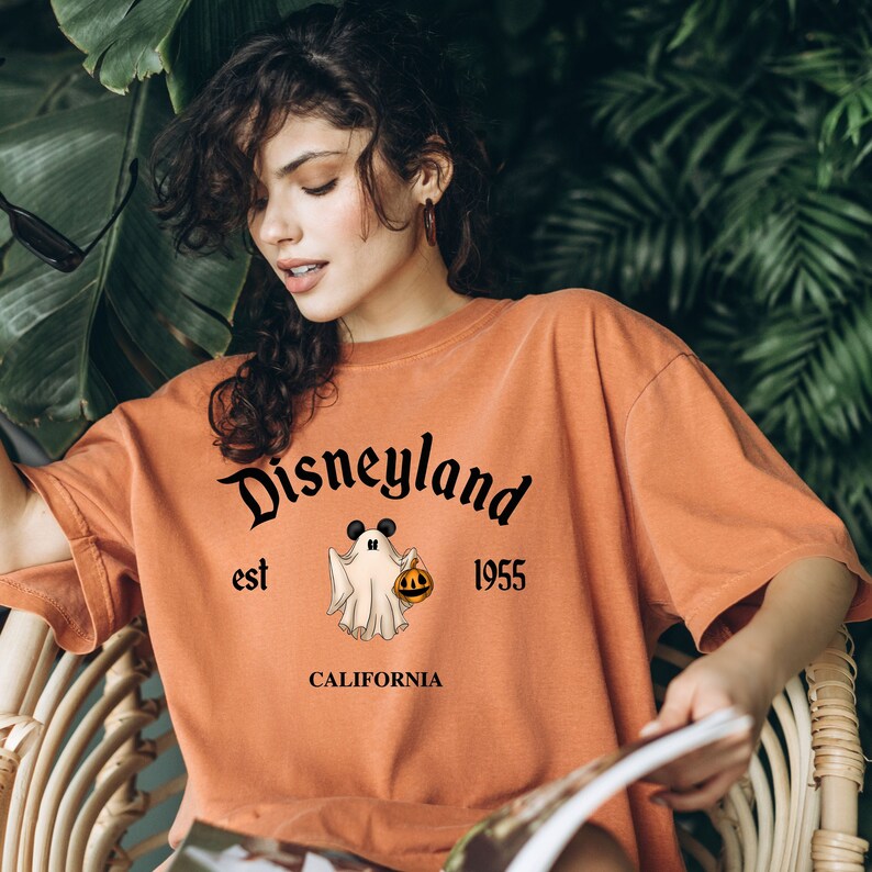 Puede incluir: Camiseta naranja con un fantasma blanco que sostiene una calabaza. La camiseta tiene el texto "Disneyland est 1955 CALIFORNIA" impreso en ella.
