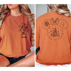 K&ouml;nnte beinhalten: Orangefarbenes T-Shirt mit einem schwarzen Motiv von Mickey Mouse und dem Text "The Happiest Place on Earth" auf der R&uuml;ckseite. Die Vorderseite des Shirts hat ein schwarzes Motiv von Minnie Mouse vor dem Schloss von Aschenputtel mit dem Text "Jessie".