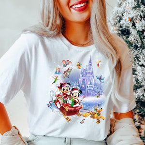 Puede incluir: Camiseta blanca con un dise&ntilde;o navide&ntilde;o colorido con Mickey Mouse y Minnie Mouse montando en un trineo con un fondo nevado. El dise&ntilde;o tambi&eacute;n incluye el castillo de Cenicienta y otros personajes de Disney.