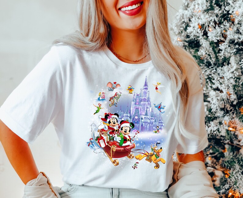 Puede incluir: Camiseta blanca con un dise&ntilde;o navide&ntilde;o colorido con Mickey Mouse y Minnie Mouse montando en un trineo con un fondo nevado. El dise&ntilde;o tambi&eacute;n incluye el castillo de Cenicienta y otros personajes de Disney.