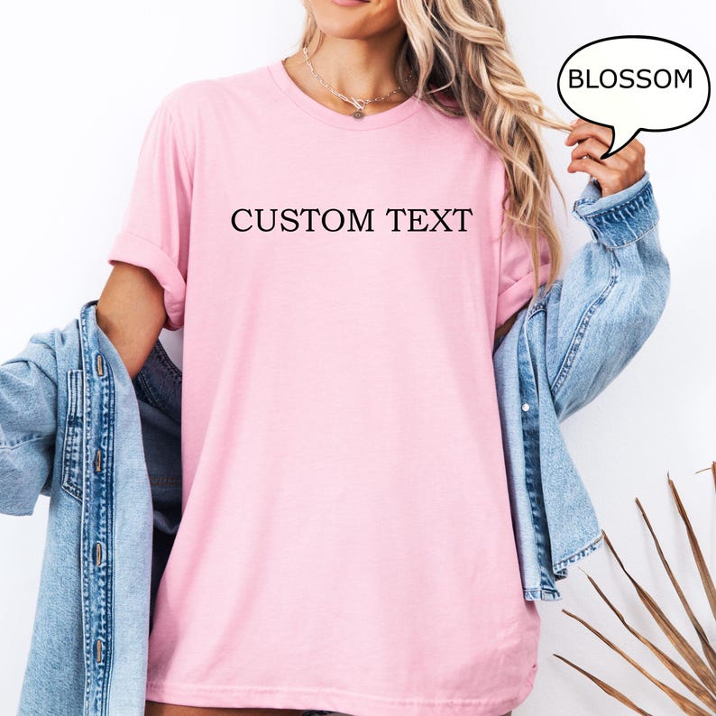 May include: A pink t-shirt with the text "CUSTOM TEXT" printed on the front. The t-shirt is being modeled by a person wearing a denim jacket. A speech bubble with the word "BLOSSOM" is in the top right corner of the image.