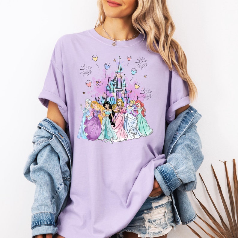 Puede incluir: Una camiseta morada con un gr&aacute;fico del castillo de Disney y siete princesas. Las princesas llevan sus vestidos caracter&iacute;sticos y caminan hacia el castillo. Hay globos y fuegos artificiales en el fondo.