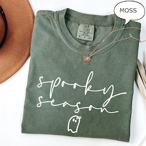 May include: A moss green t-shirt with the words "spooky season" in white script, and a ghost graphic. A gold necklace with a pendant is draped over the shirt. The shirt is folded on a white surface.