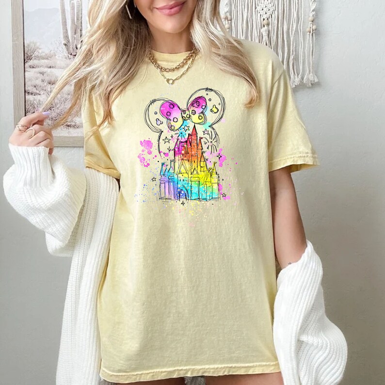 Puede incluir: Una camiseta amarilla con un boceto de un castillo y orejas de Mickey Mouse en colores arco&iacute;ris. El castillo est&aacute; rodeado de estrellas y salpicaduras de pintura.