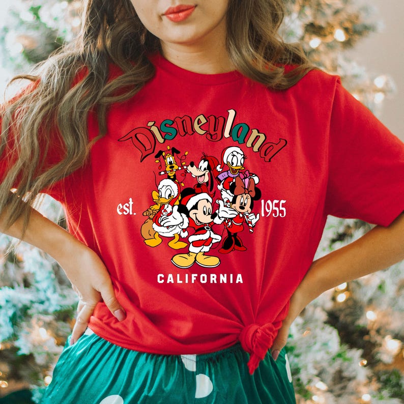 May include: Red t-shirt with a Christmas-themed Disneyland graphic featuring Mickey Mouse, Minnie Mouse, Donald Duck, Goofy, Pluto, and a Christmas tree. The text reads "Disneyland est. 1955 California".