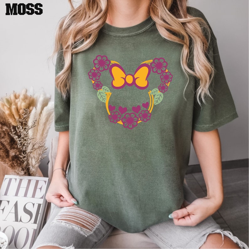 May include: A green t-shirt with a floral design featuring a yellow bow. The design is in the shape of a heart.